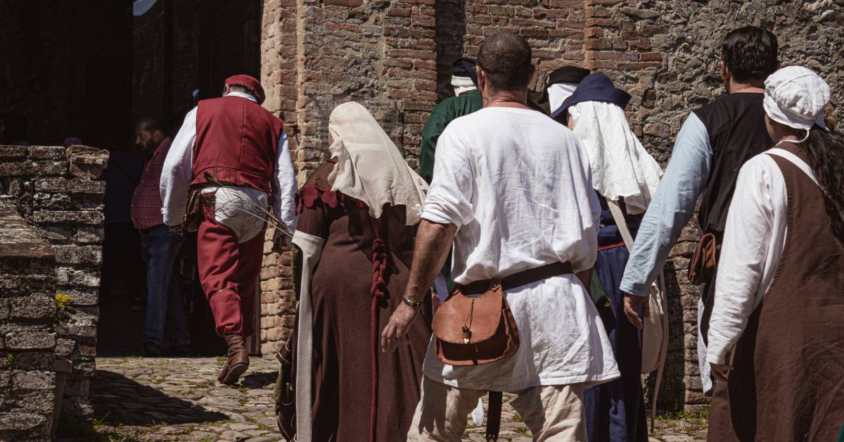 Group of unrecognizable people dressed in medieval costumes walking down an ancient street leading to a castle.
