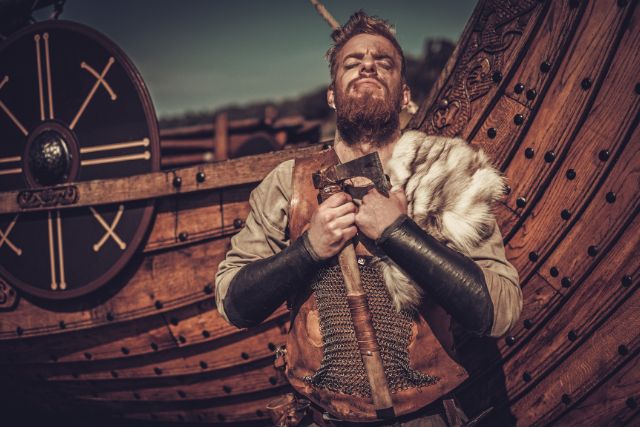 A viking warrior holding an axe with a ship behind it.