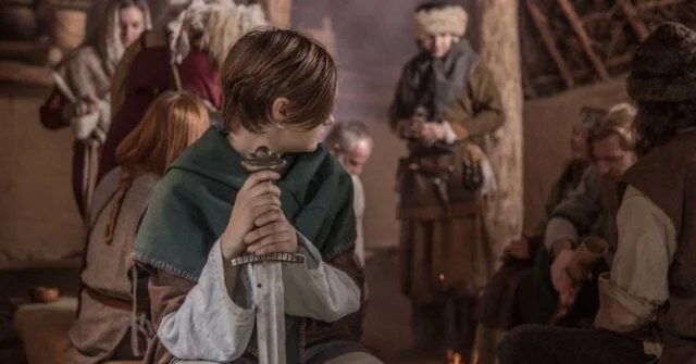 A viking boy holding a sword in village settlement gathering.