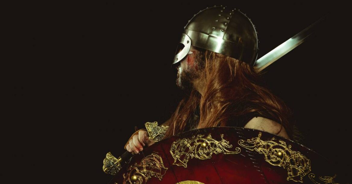 A viking warrior in side view wearing full battle gear and holding a sword and shield.