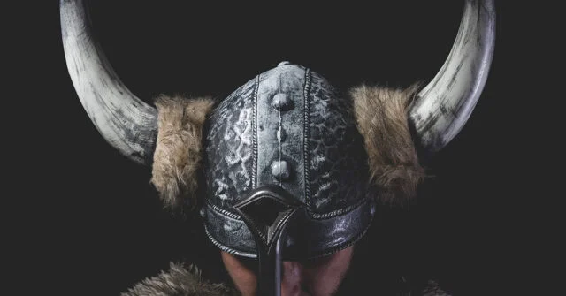 Viking warrior wearing a horned helmet bowing head resting on his sword.